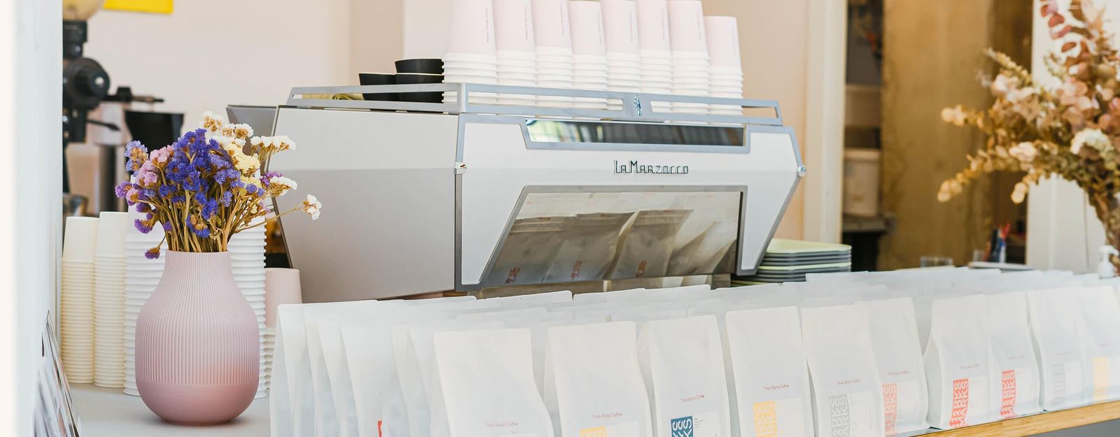 Biała KB90 na barze kawiarni, obok kwiaty i torebki kawy